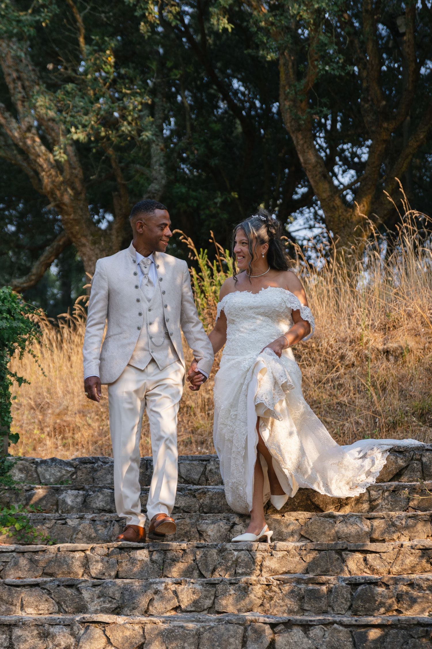 Newlyweds in white attire pose on stone wall during golden hour photo session.
