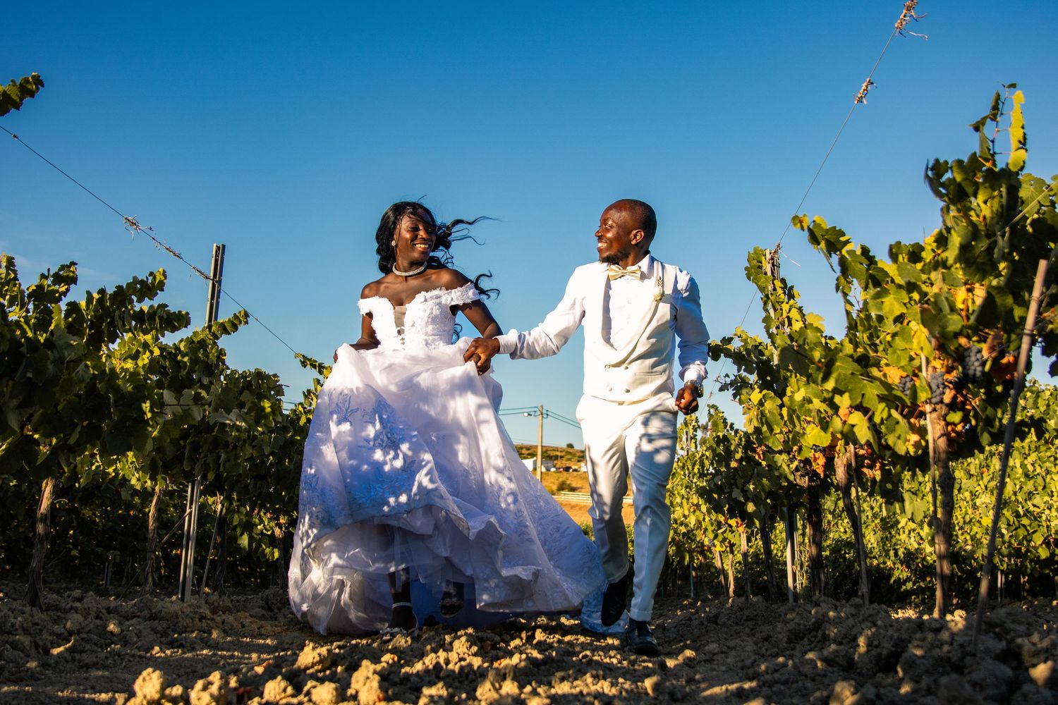 A couple in white attire dances joyfully in a sunlit vineyard during their wedding celebration.
