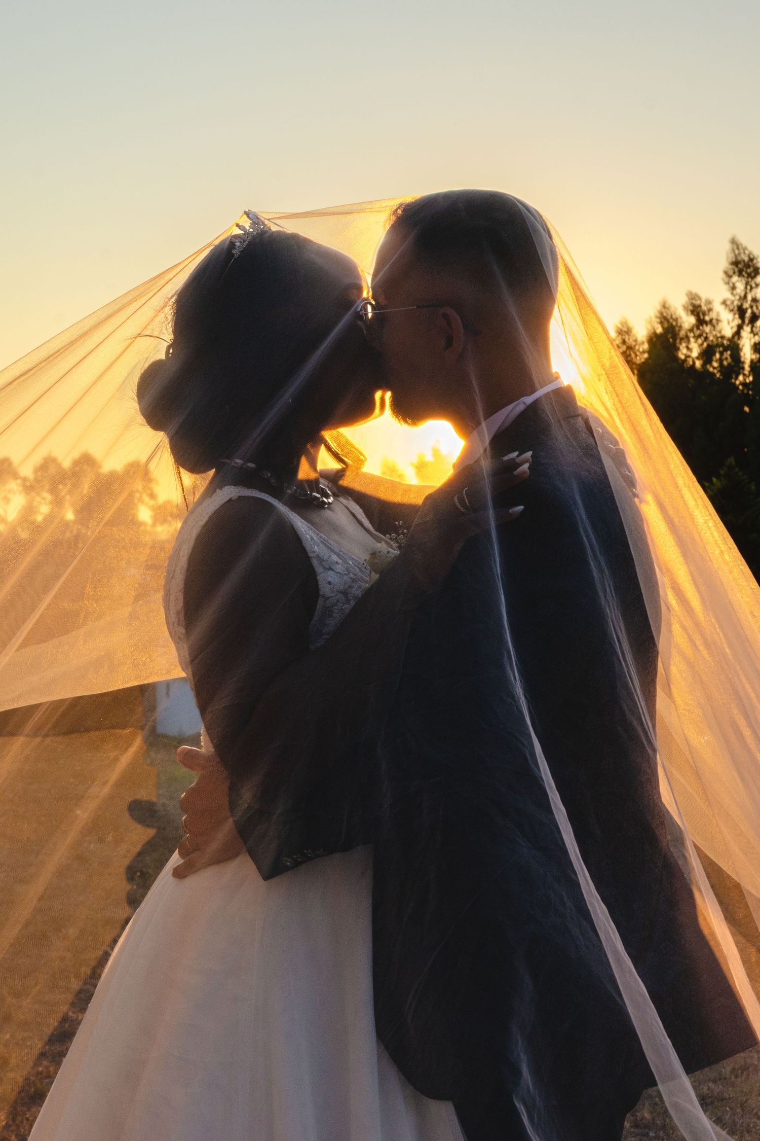 Romantic sunset wedding photo with flowing veil and silhouetted couple embracing.