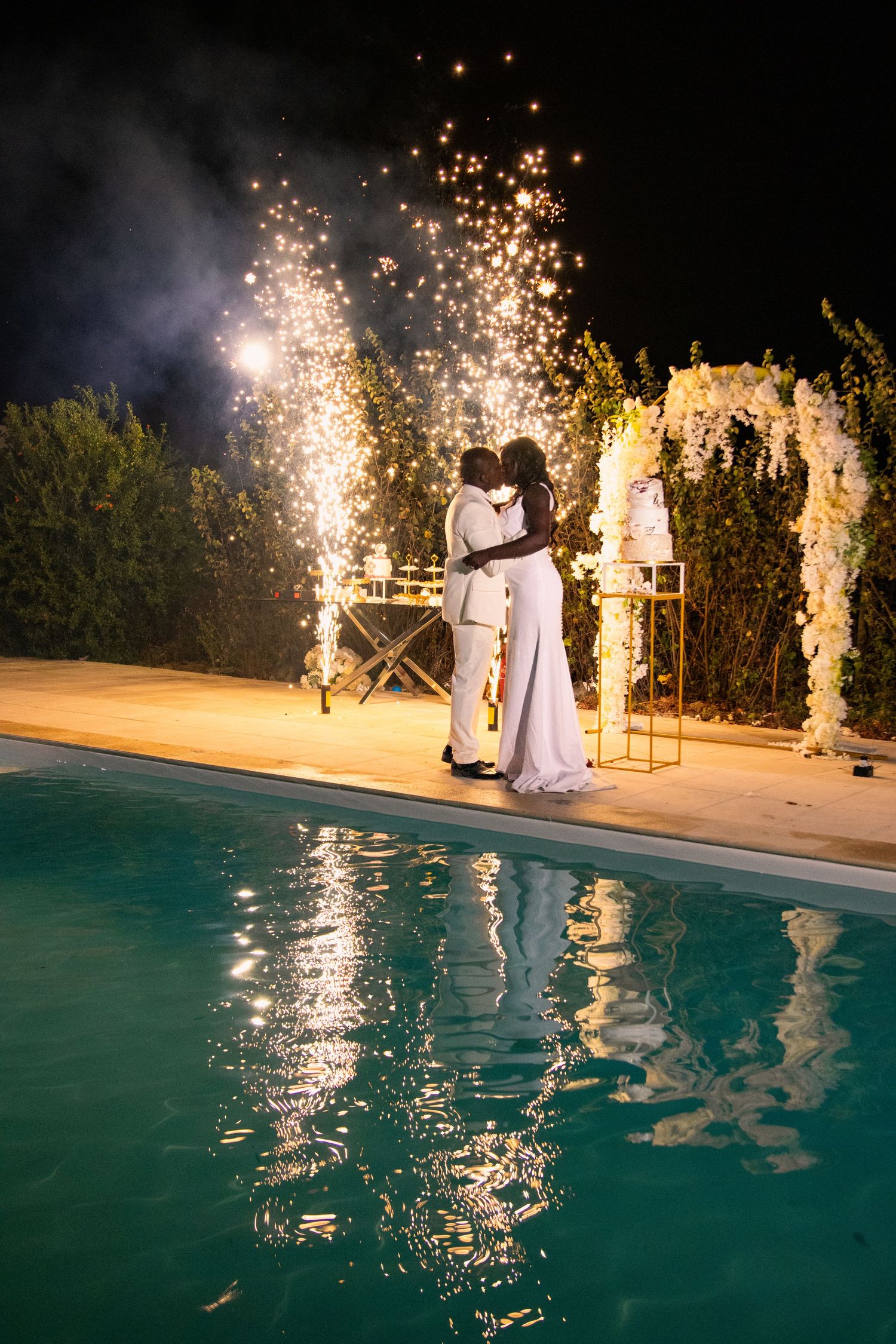 Sparklers illuminate a nighttime wedding celebration by a turquoise pool.