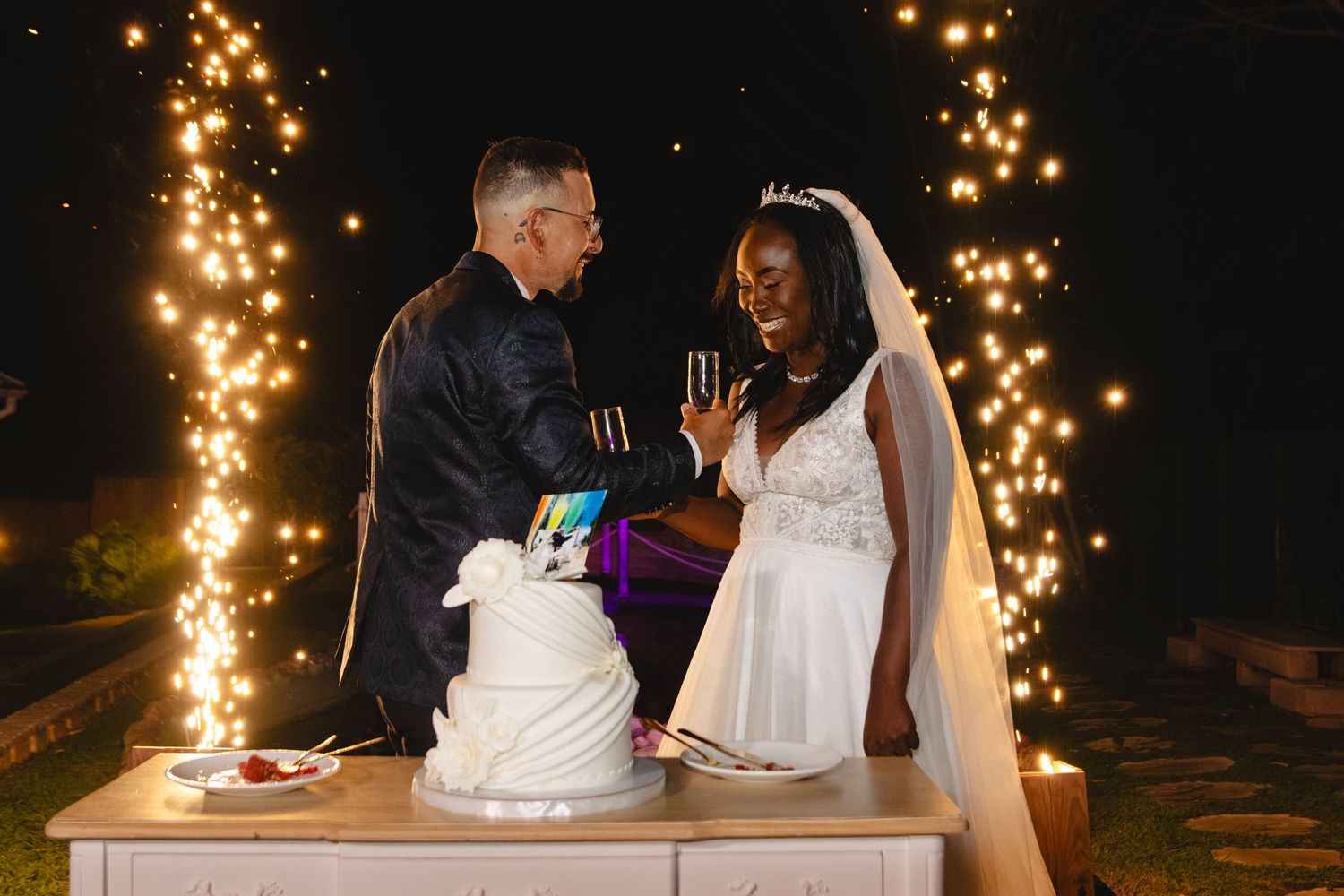 Newlyweds cut their wedding cake together surrounded by sparkling lights.