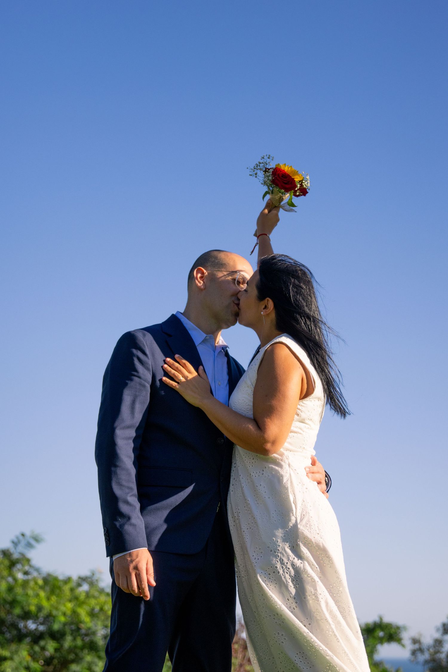 Newlyweds embrace against a bright blue sky during their outdoor wedding celebration.