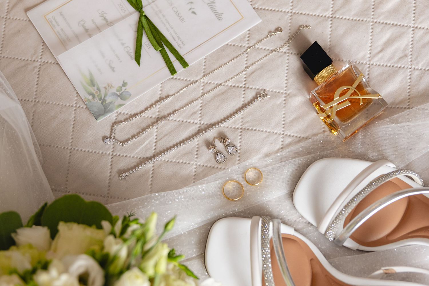 Wedding accessories including rings, perfume, and invitations arranged on white fabric.