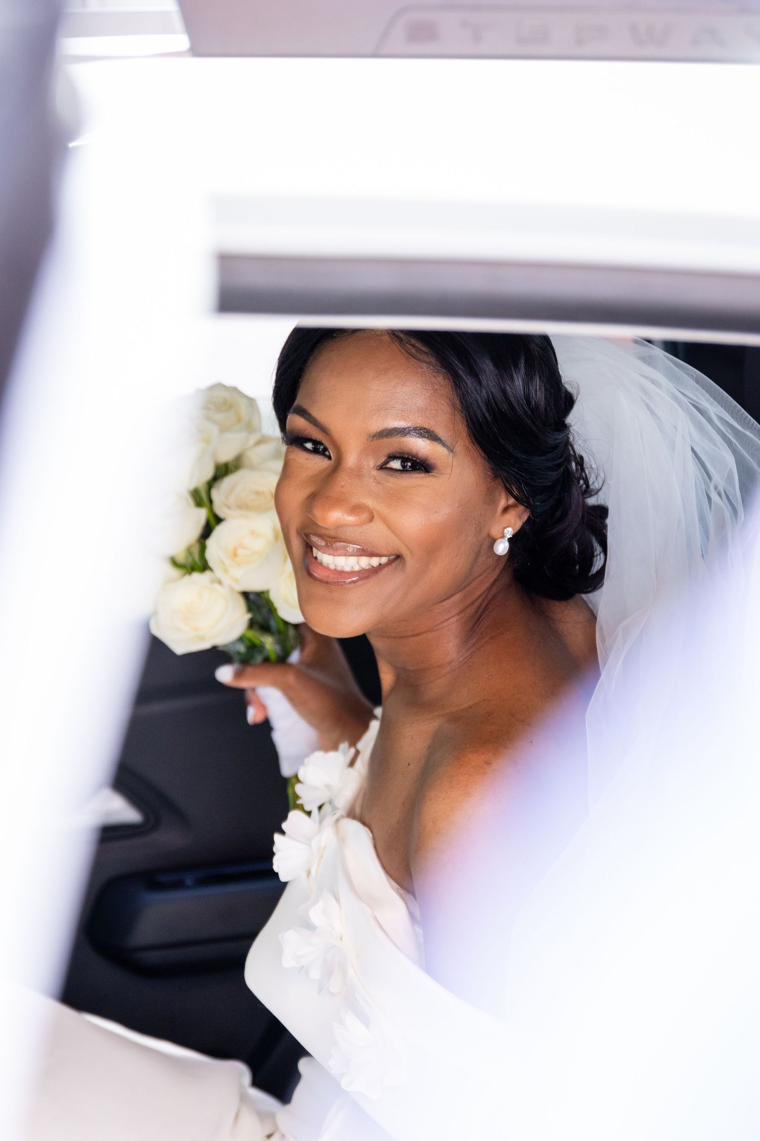 Radiant bride in white dress and veil holding bouquet inside wedding car.