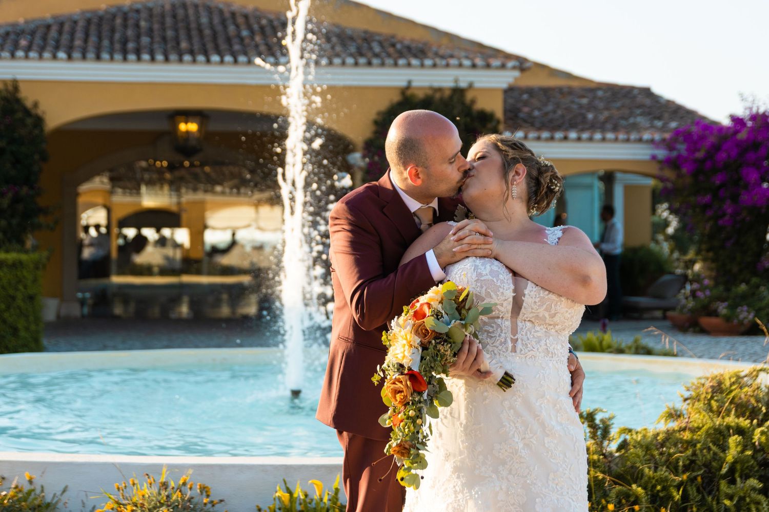 A couple shares a kiss by a fountain with sunflowers in a garden setting.