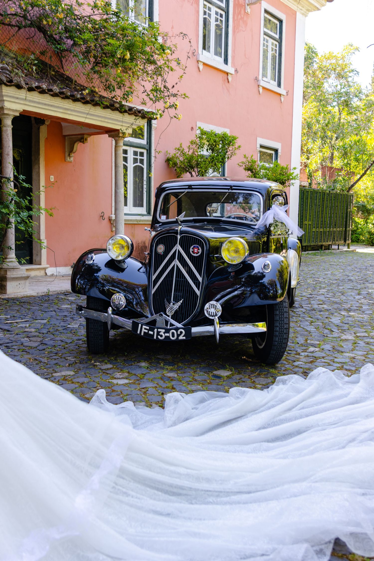Classic vintage car parked outside pink building with flowing white wedding dress train.