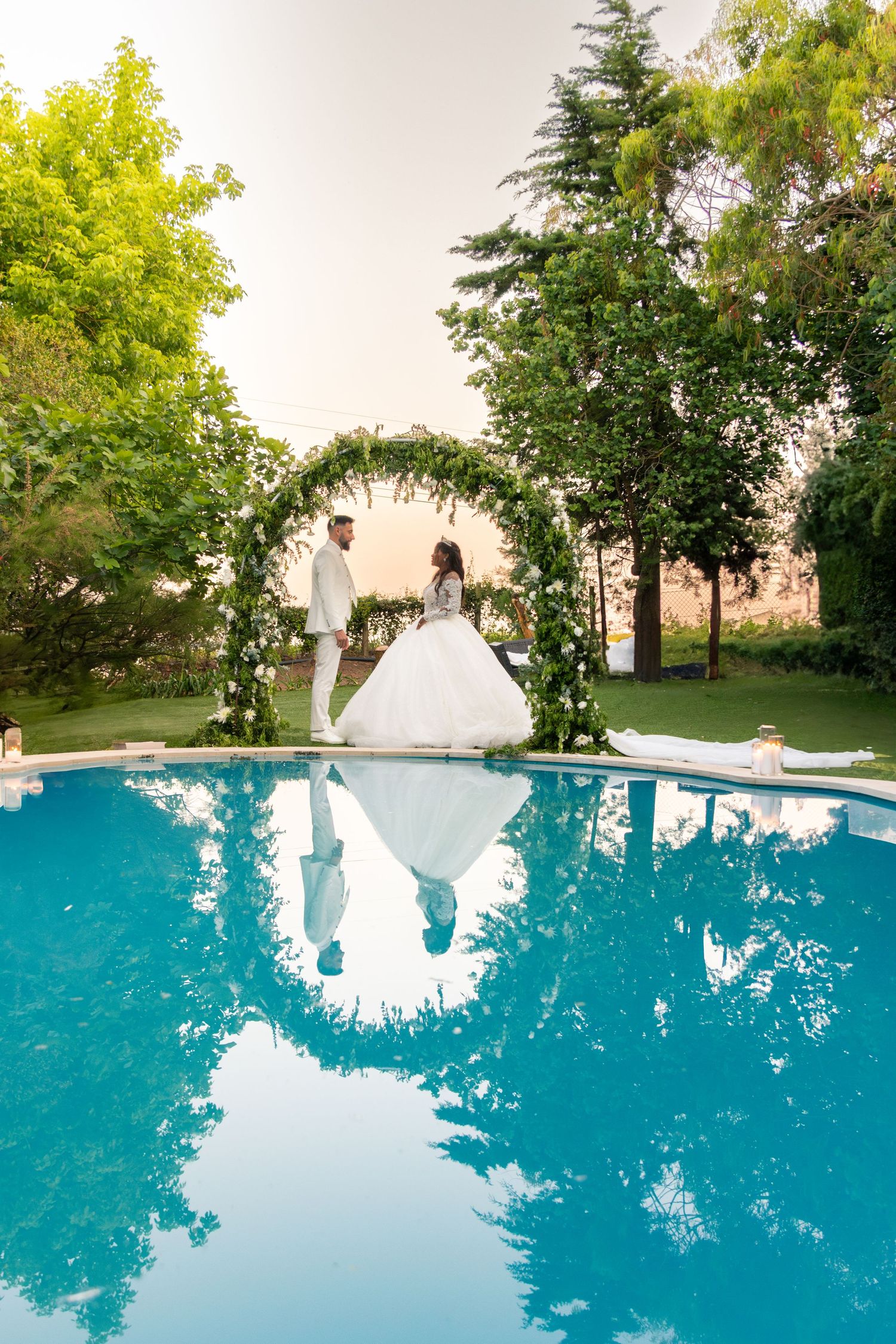 Wedding couple's reflection captured in a serene blue pool surrounded by trees.