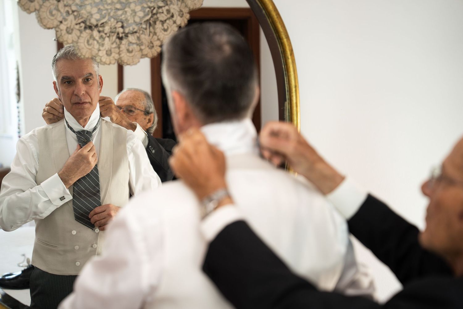 Man adjusting tie while getting ready in front of ornate mirror during wedding preparations.