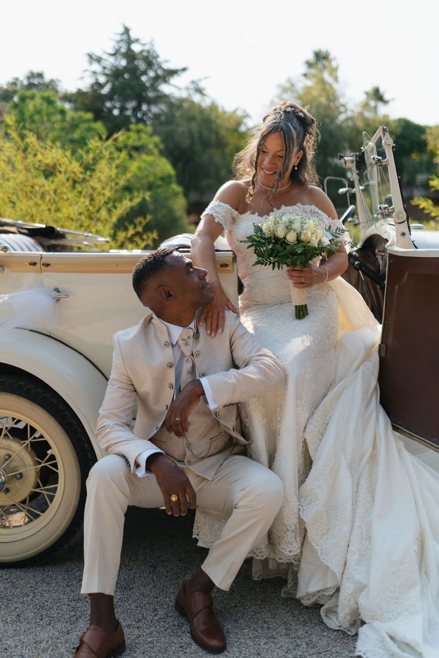 Playful wedding couple poses by vintage white car during outdoor photo session.