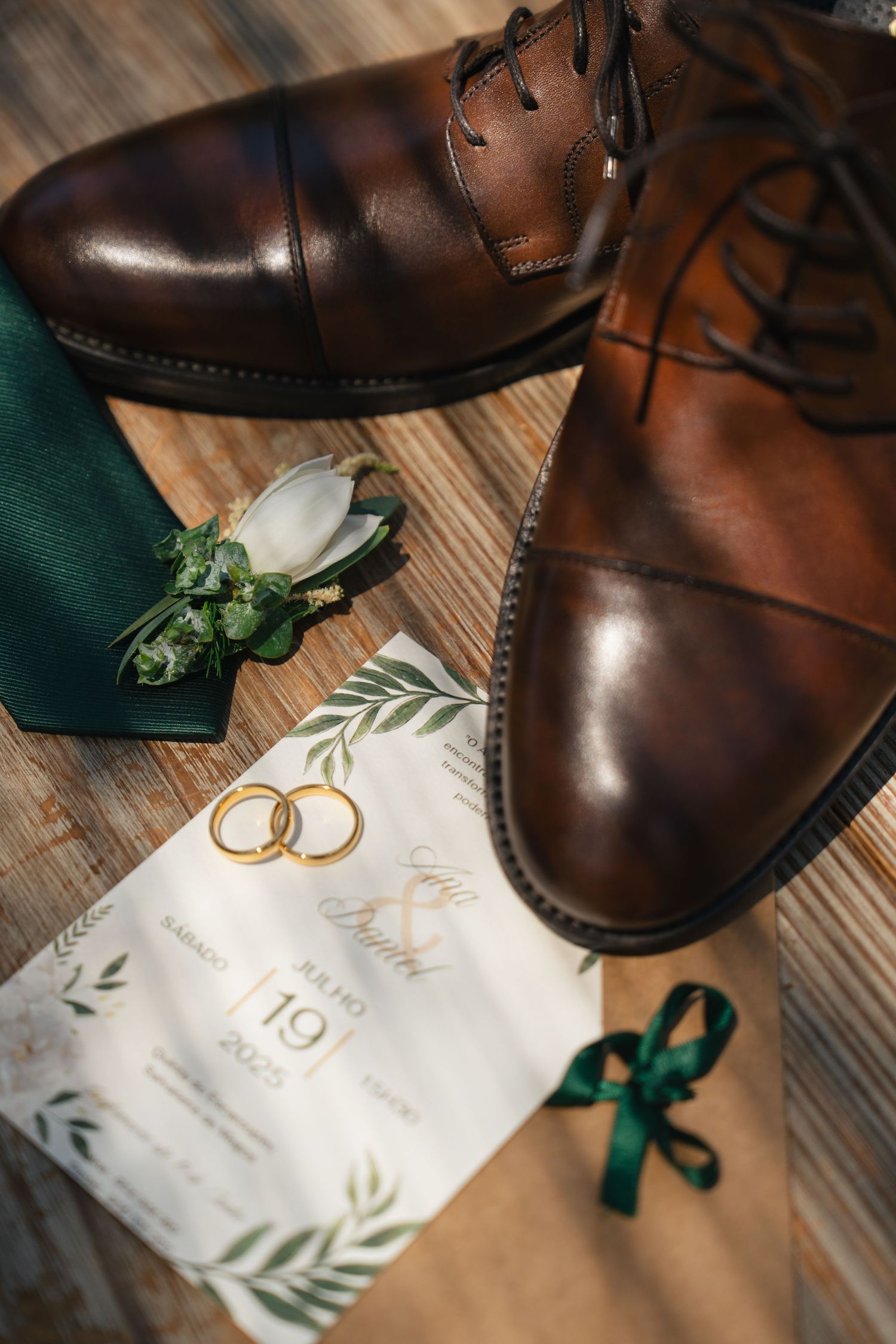 Brown leather dress shoes and wedding rings displayed with an elegant invitation featuring greenery.
