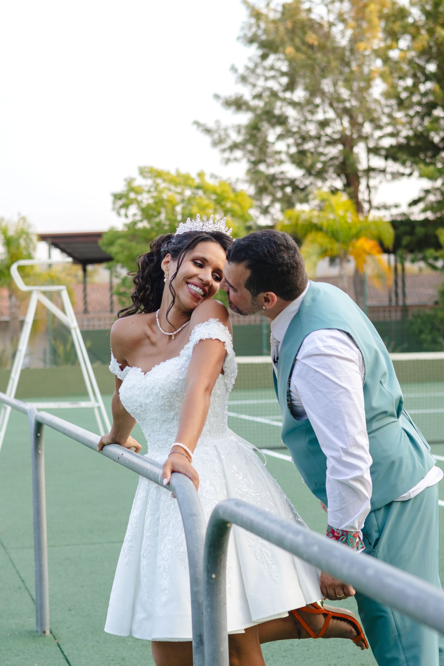 A couple sharing a playful moment on a tennis court, with the bride wearing a short white dress.