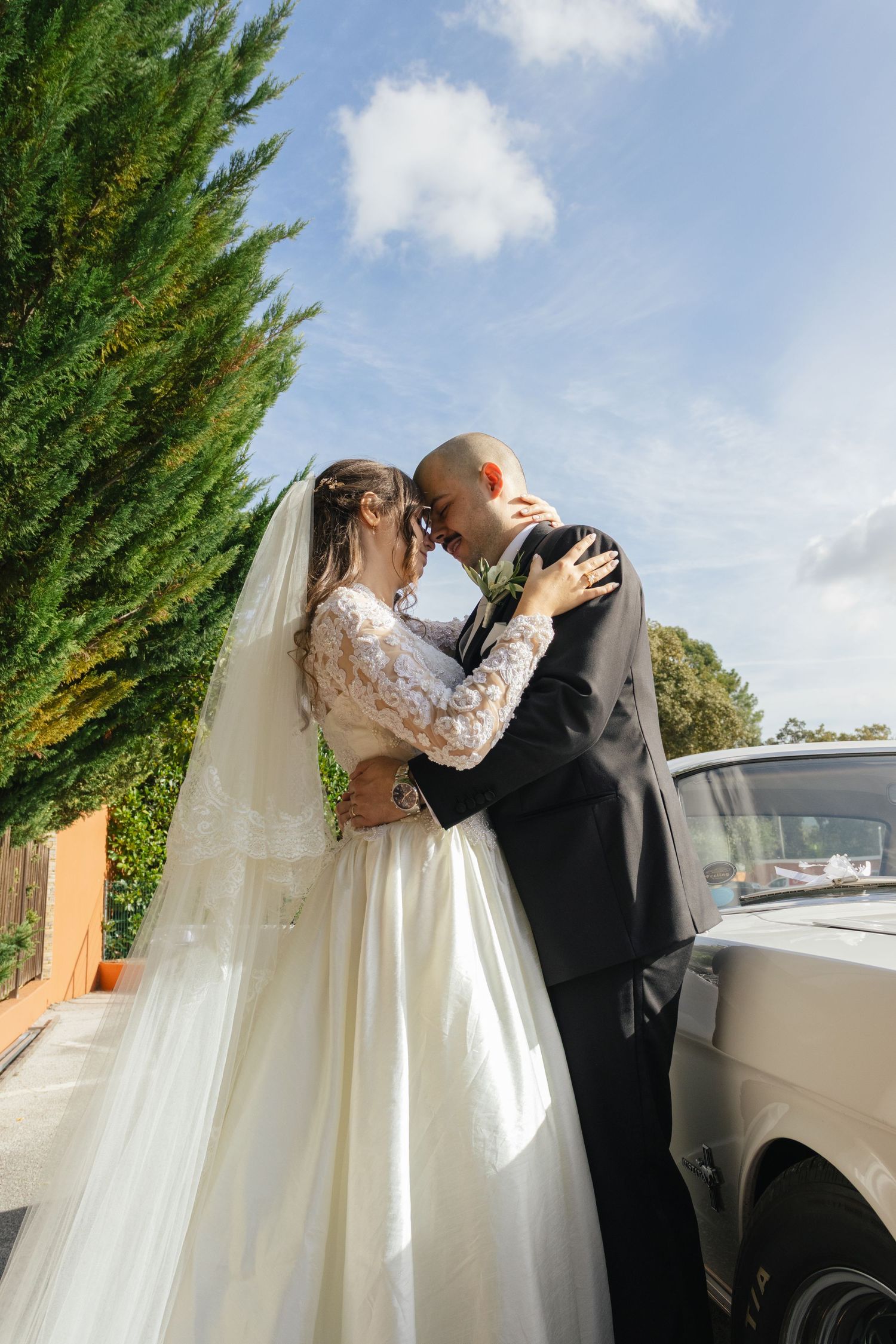 Newlyweds share an intimate moment beside a vintage car under blue skies and cypress trees.