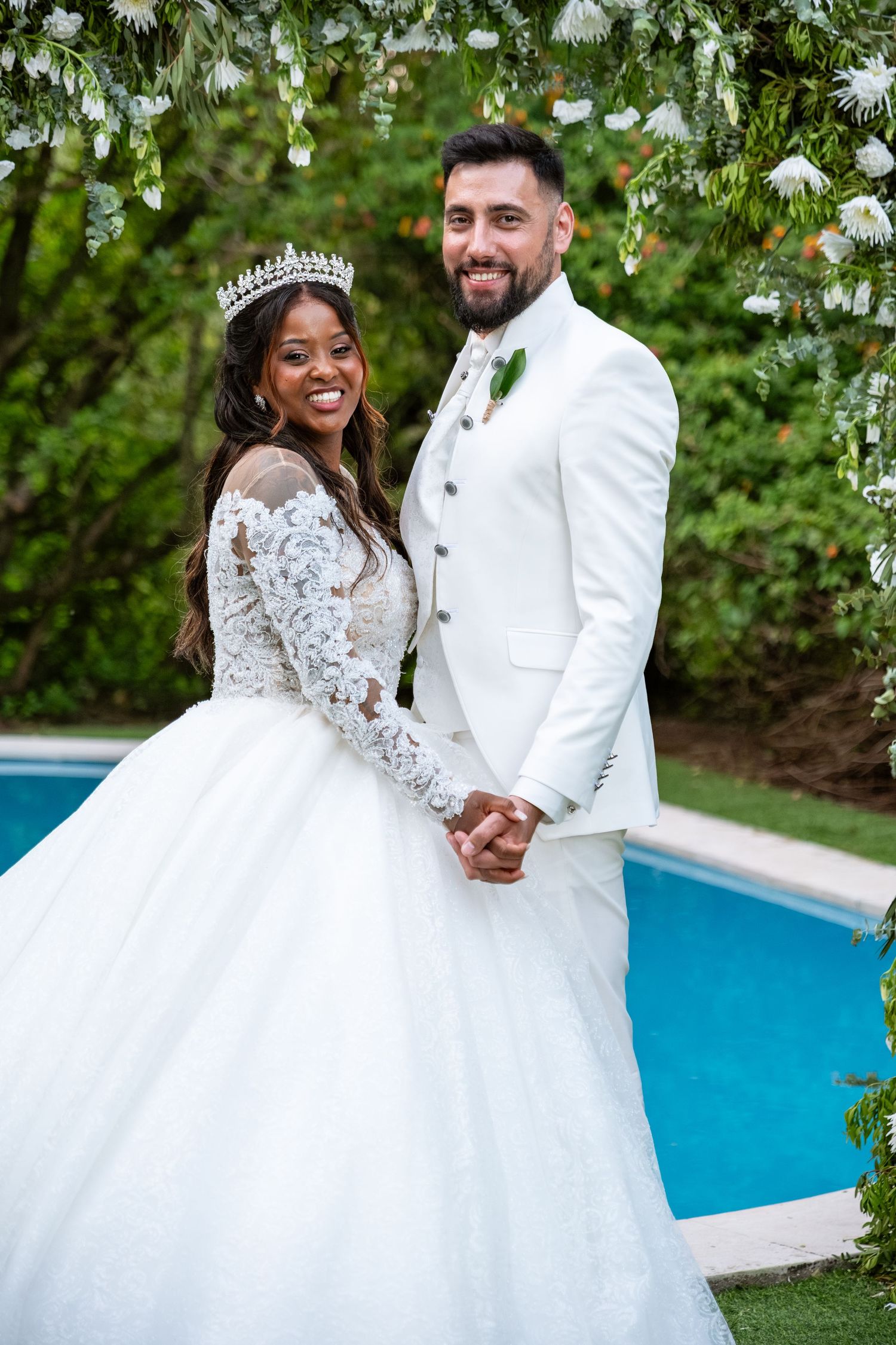 A couple in elegant white wedding attire poses by a swimming pool surrounded by lush greenery.