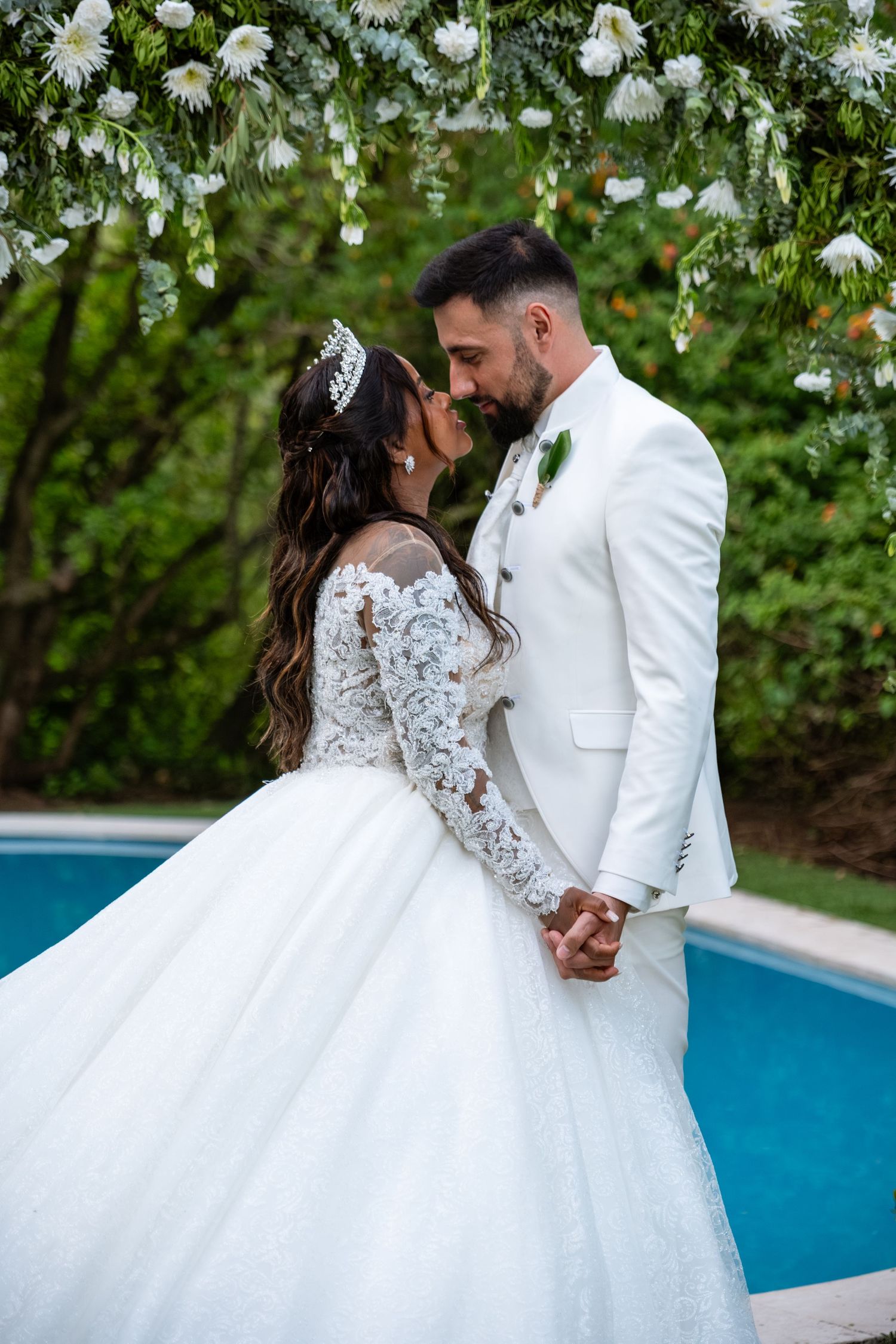 Newlyweds share a romantic moment in white attire near a bright blue pool.