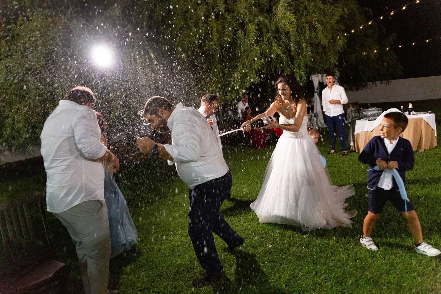 Wedding guests dance under twinkling lights at an outdoor evening reception.