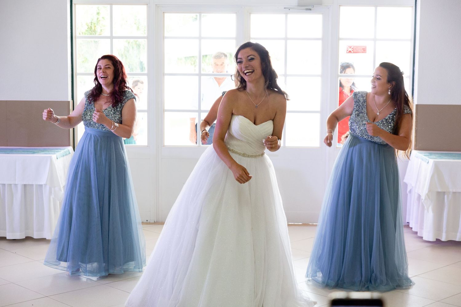 Bridesmaids in light blue tulle skirts dance and celebrate in a bright indoor venue.
