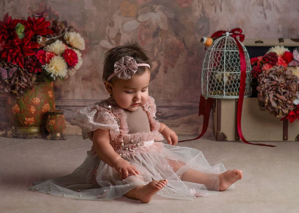 Baby in pink tulle dress sits on floor surrounded by vintage decor and red floral arrangements.