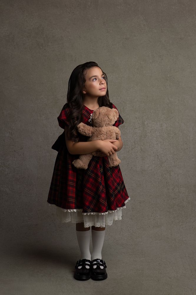 Full length studio portrait with teddy bear and plaid dress against neutral background.