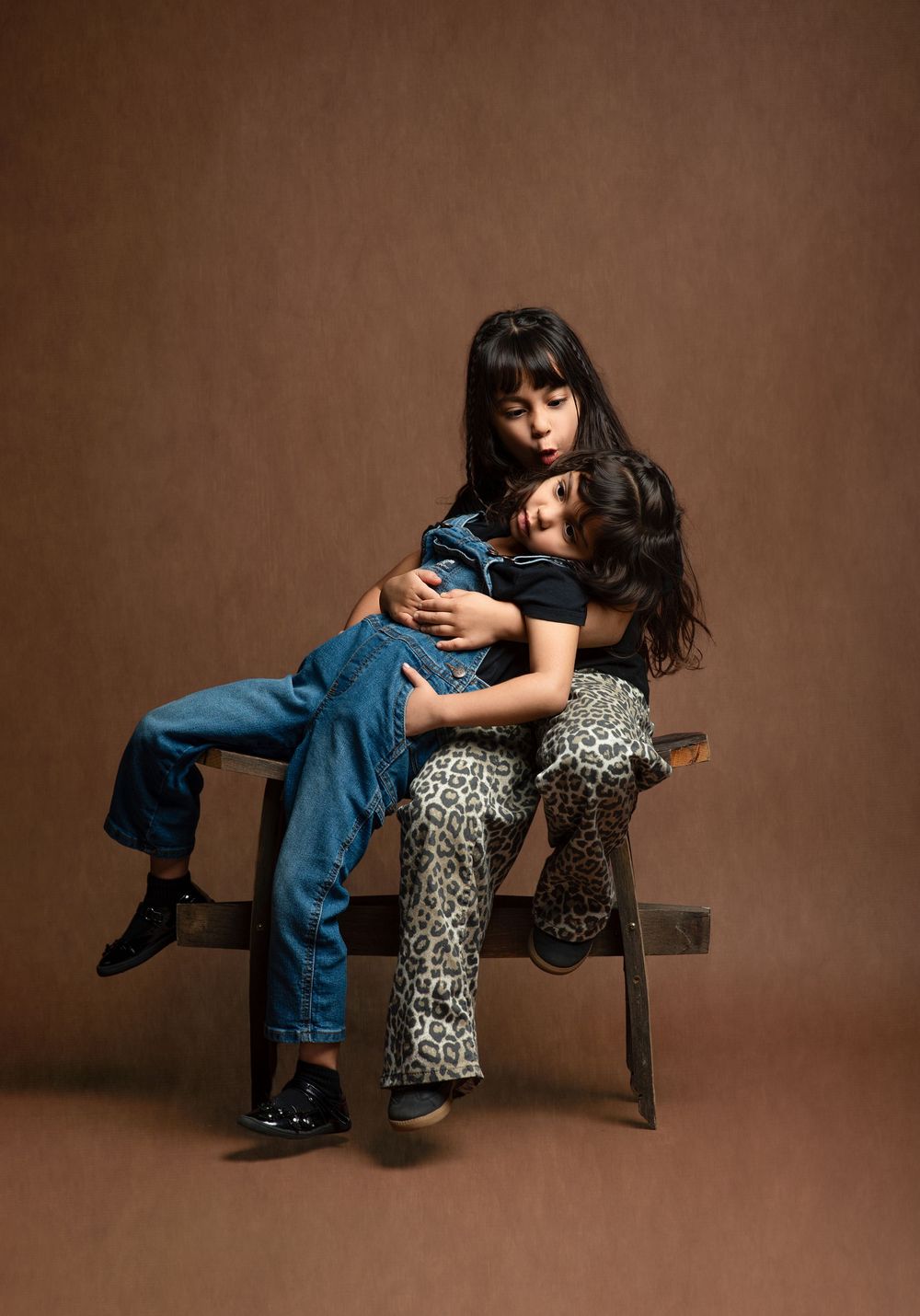 Two people in contrasting outfits of denim and leopard print pose intimately on a wooden chair against brown backdrop.