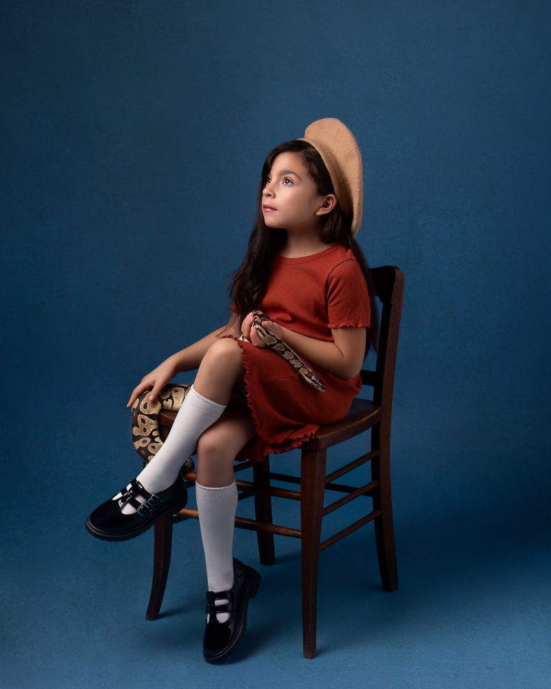 Studio portrait on wooden chair wearing red outfit and beige hat against blue background.