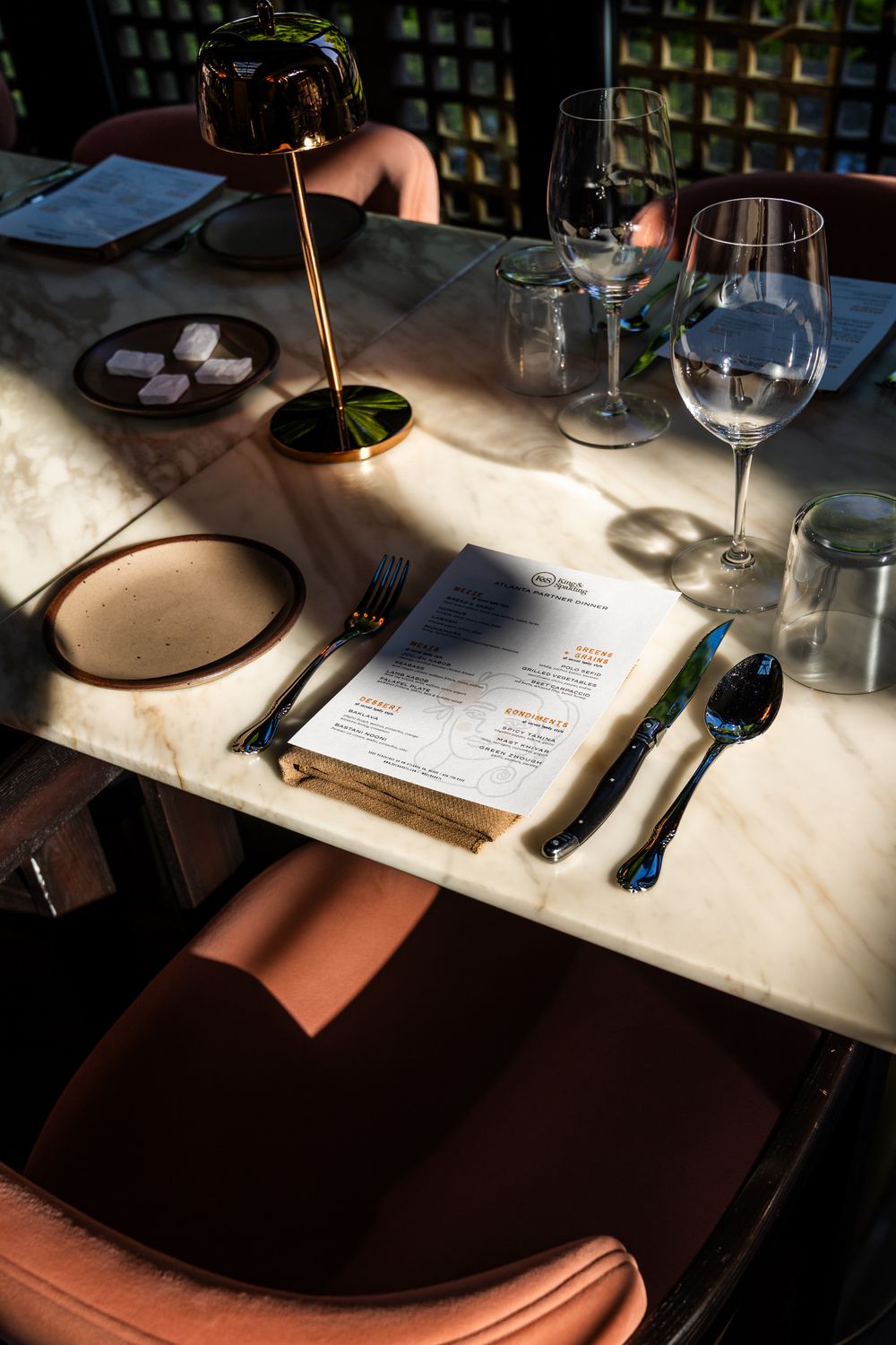 Upscale restaurant place setting with menu, wine glasses, and brass lamp on marble table.