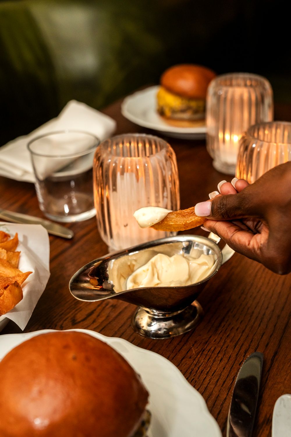 Candlelit dinner table with hamburgers and chips while someone dips into a creamy sauce in a silver bowl.