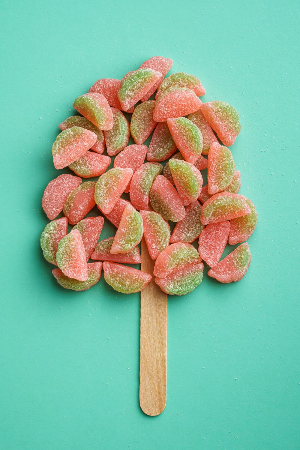 Watermelon-shaped gummy candies arranged in a tree shape on a wooden popsicle stick against turquoise background.