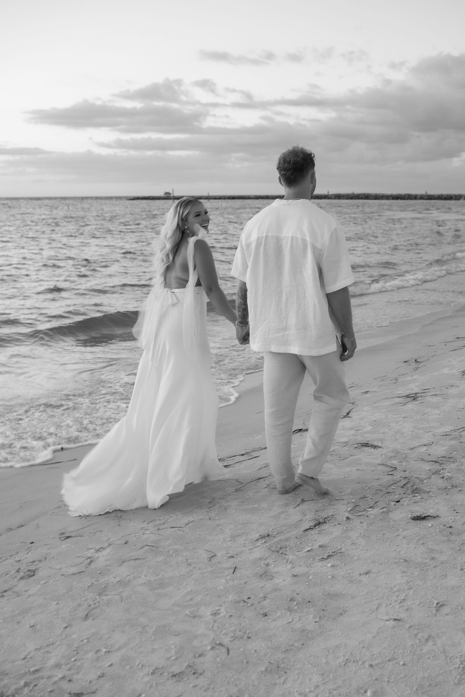 A couple walking hand in hand along the shoreline at sunset in flowing white attire in Clearwater, Florida