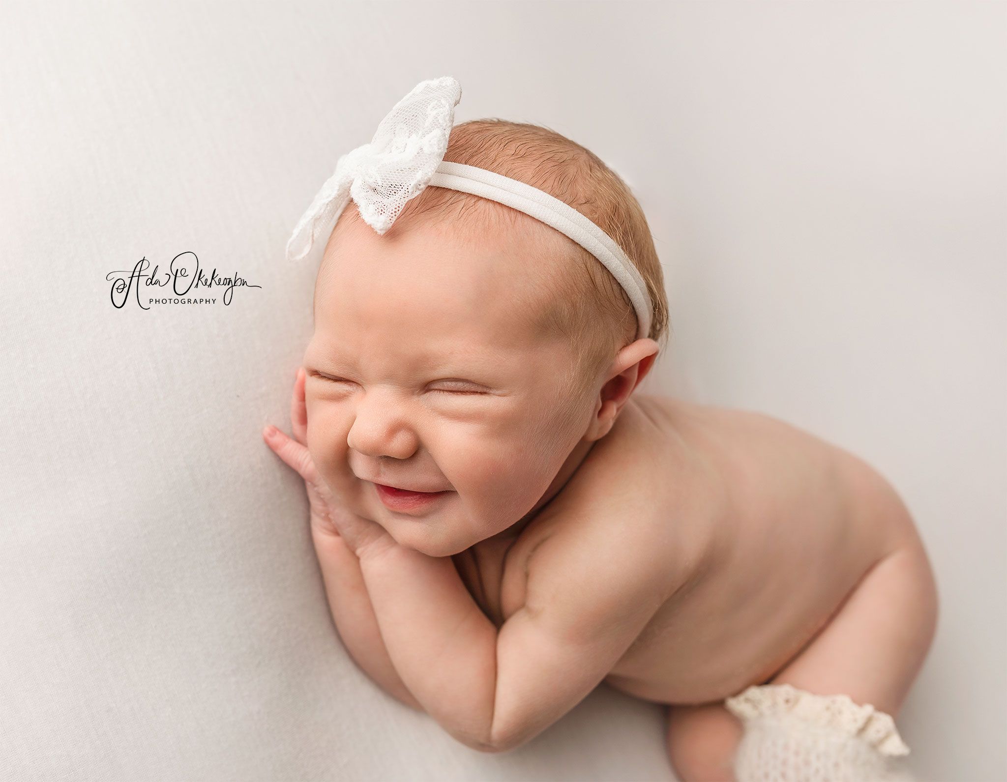 Newborn baby wearing white bow headband smiles while laying on side during professional photo session.