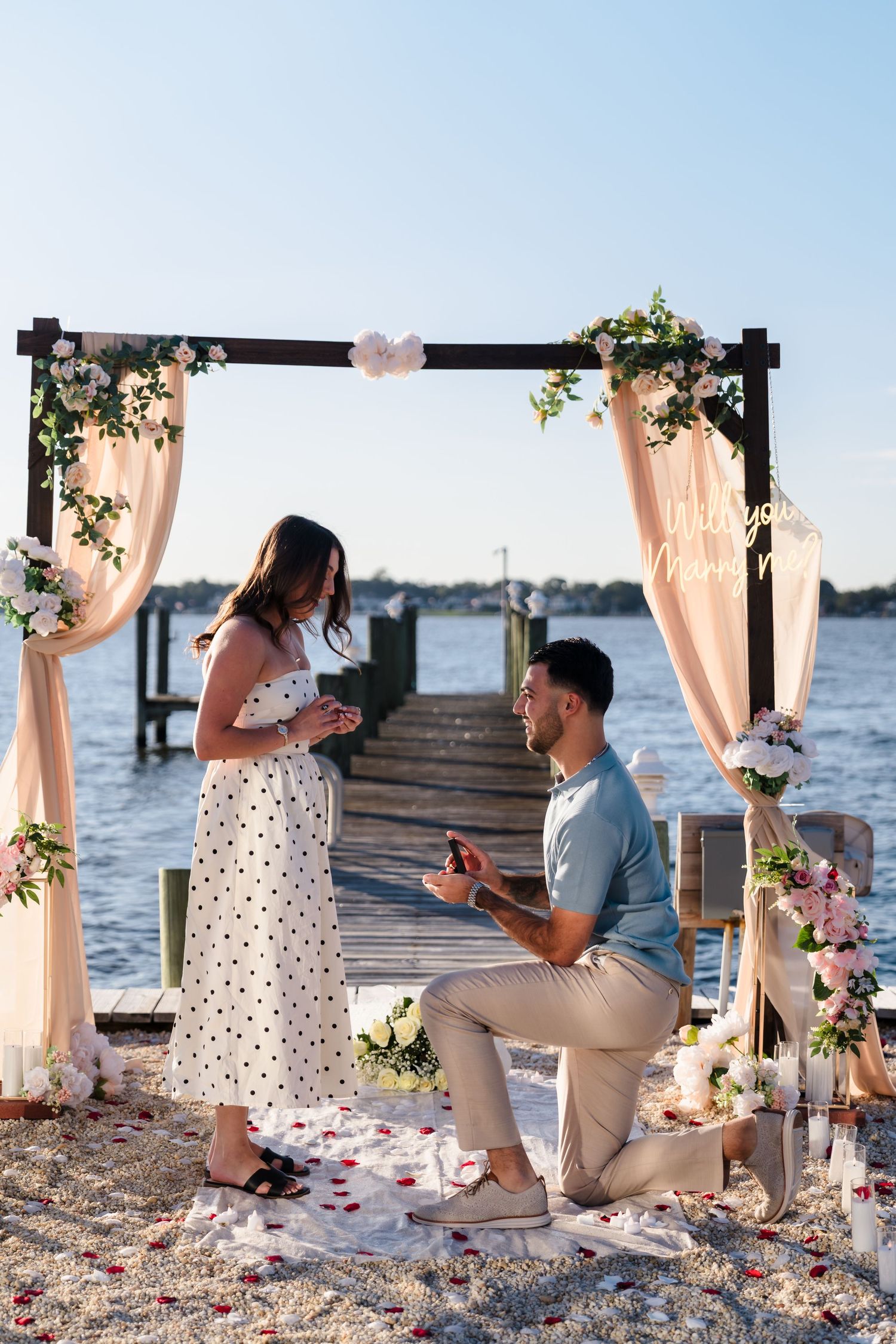 Romantic beach proposal setup with decorated arch, rose petals, and flowing curtains at sunset.