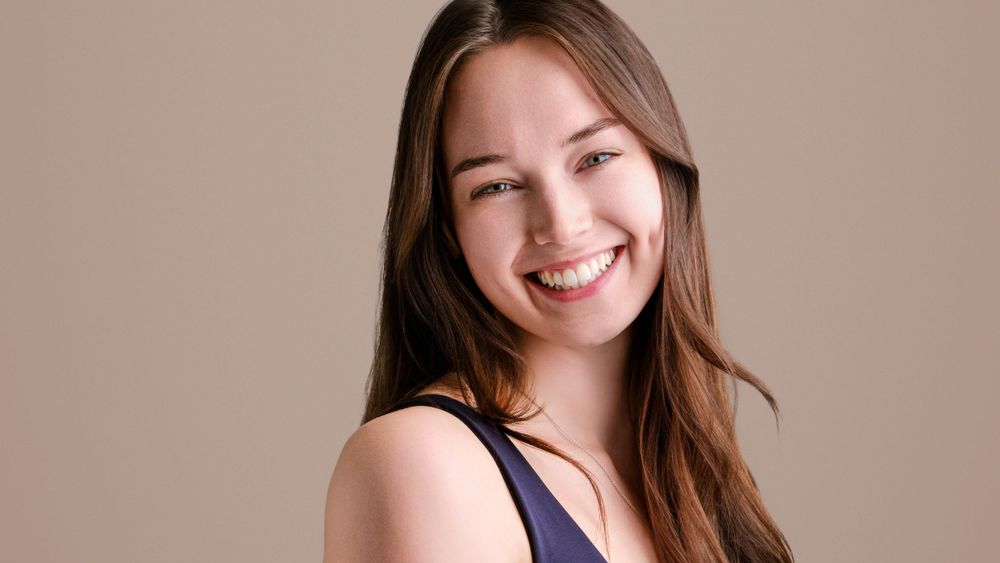 Smiling young woman with long brown hair wearing a navy top on a beige background.