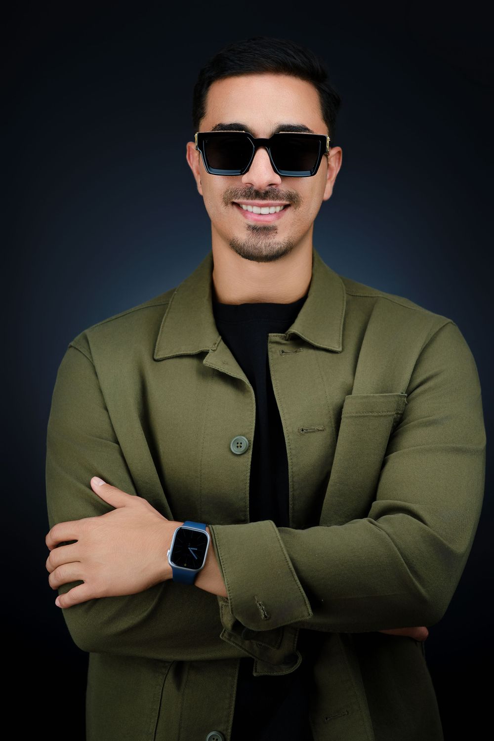 Stylish young man wearing sunglasses and olive jacket with arms crossed and smartwatch on dark background.