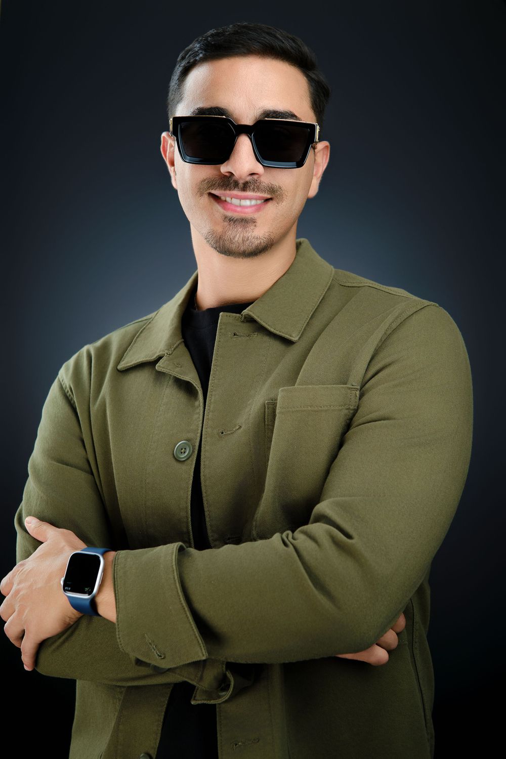 Smiling man in green jacket and black sunglasses posing with arms crossed on dark background.