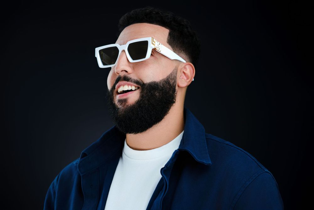 Bearded man smiling in white square sunglasses and navy shirt against dark background.