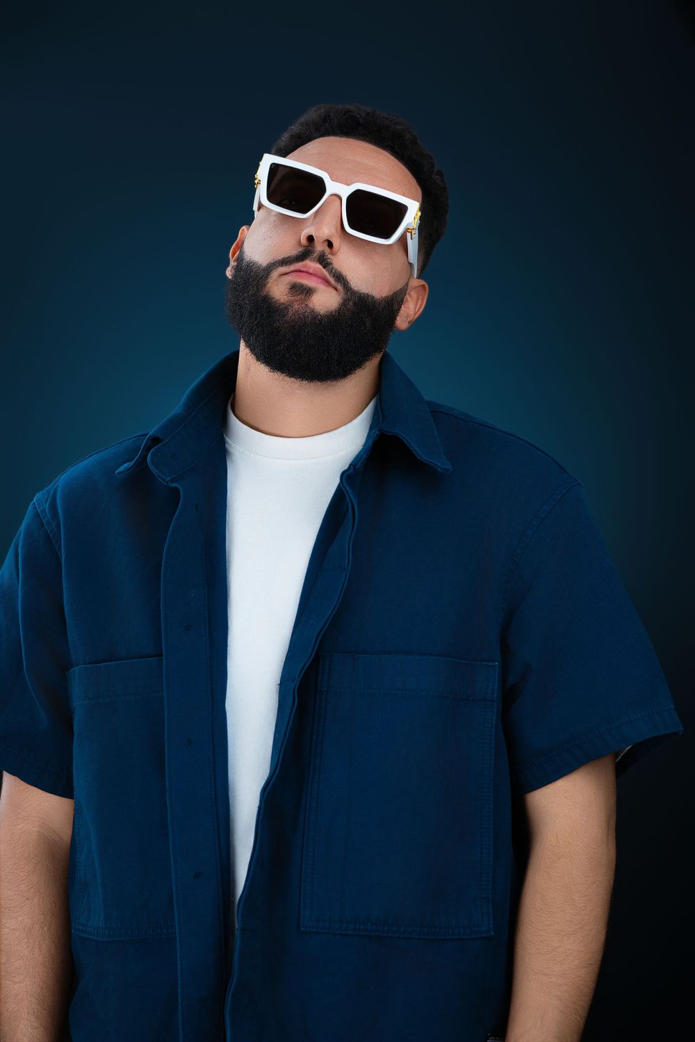 Bearded man in navy blue shirt wearing white sunglasses posing confidently on dark teal background.