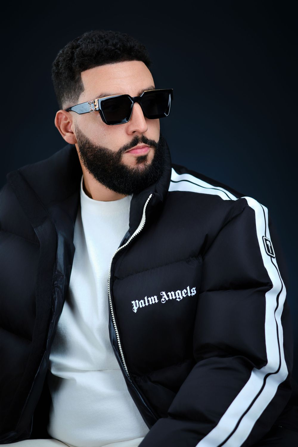 Bearded man in black Palm Angels puffer jacket and sunglasses posing on dark background.