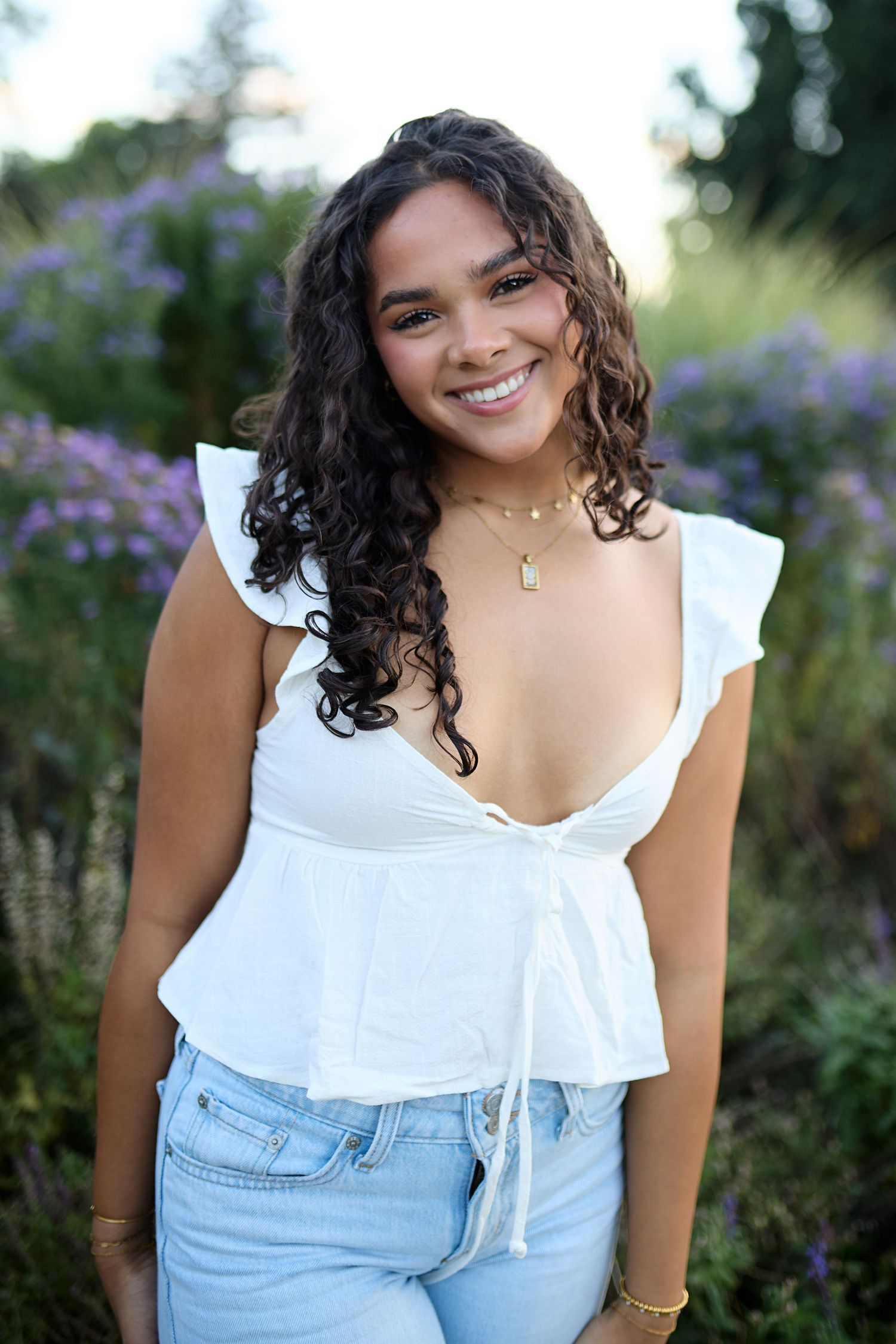 Woman in white ruffled top and denim overalls poses in garden with purple flowers in background.