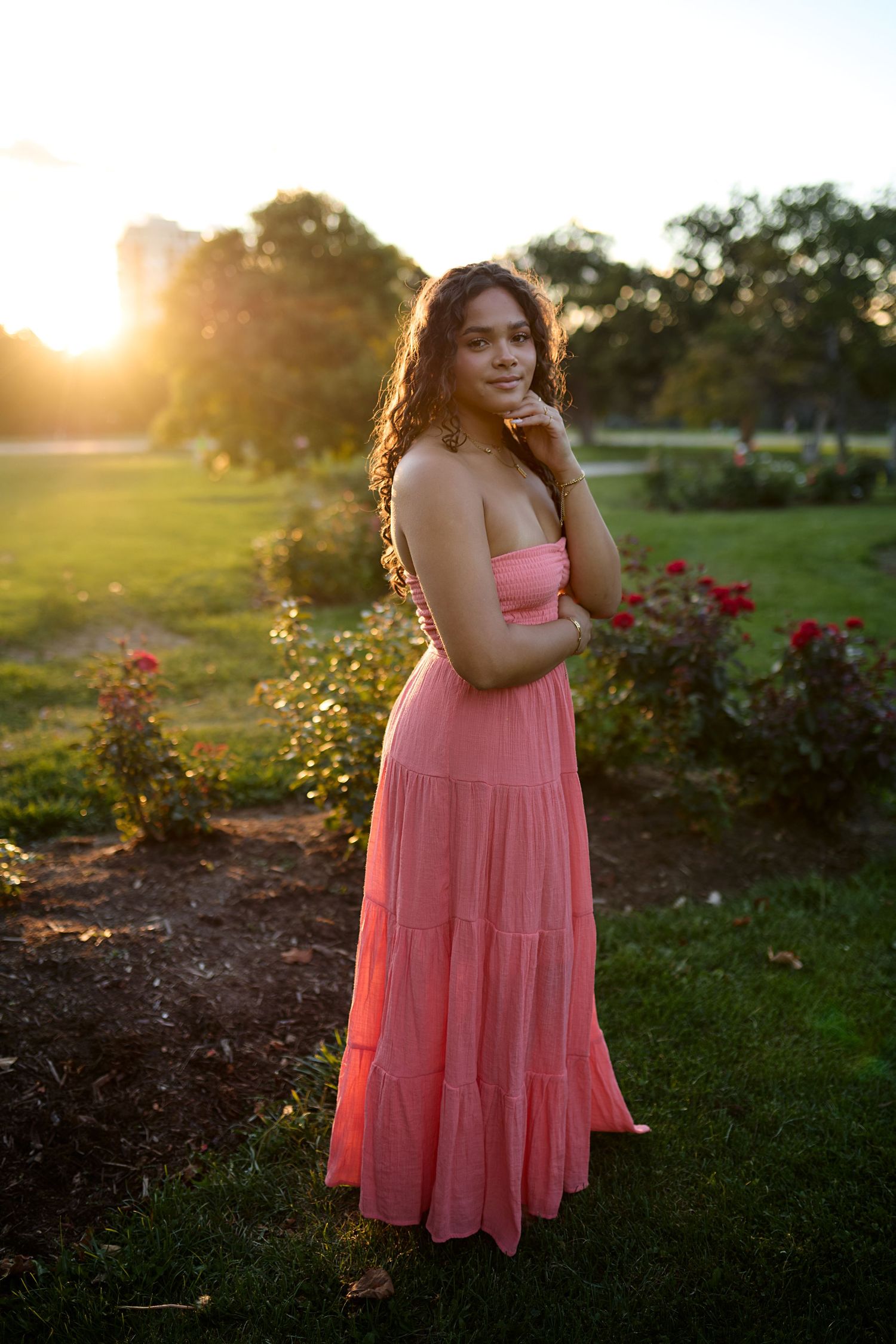 A figure in a flowing pink summer dress poses gracefully in a garden with roses at golden sunset.