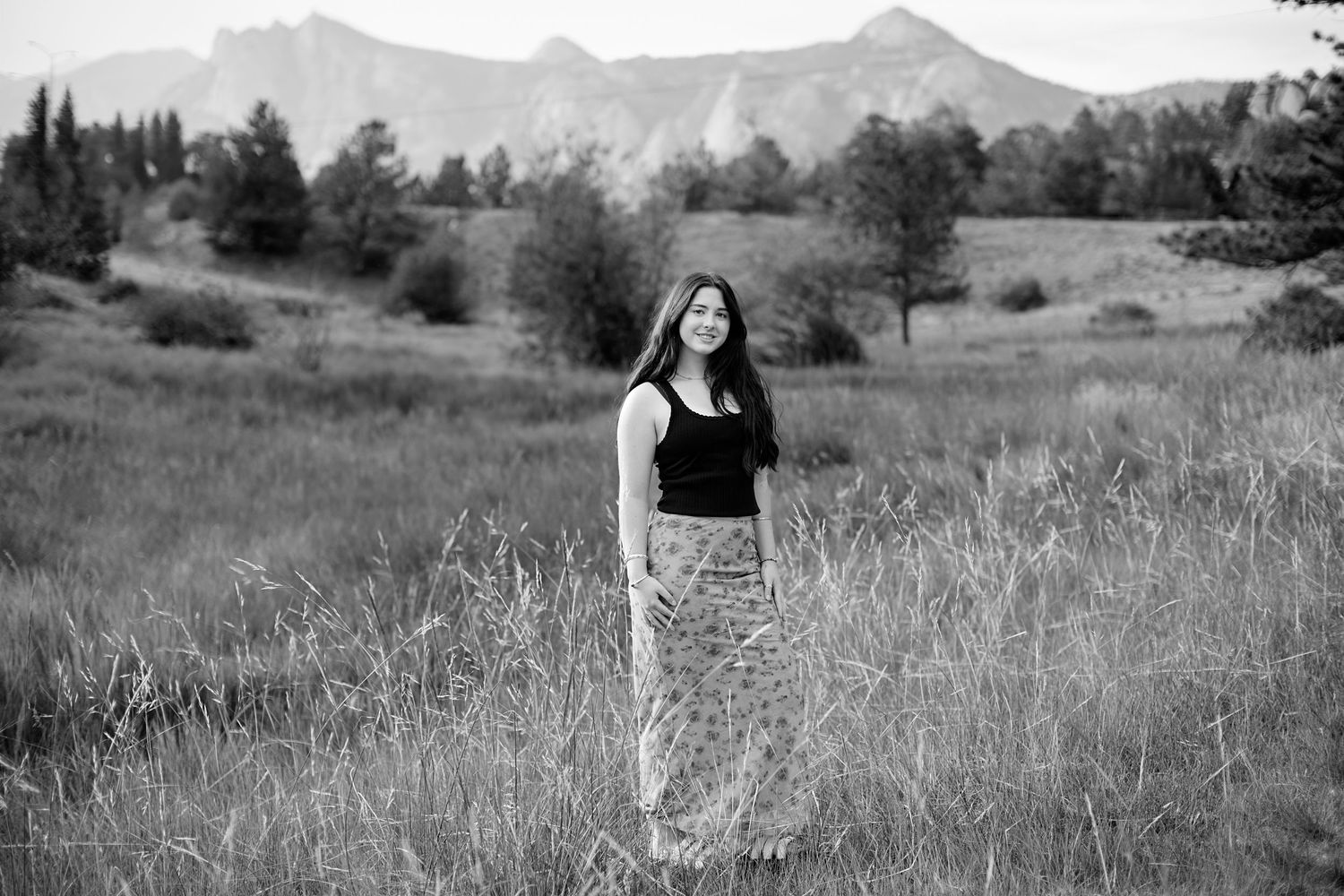 A figure stands in a grassy meadow wearing a black top and patterned skirt with mountains in the distance in black and white.