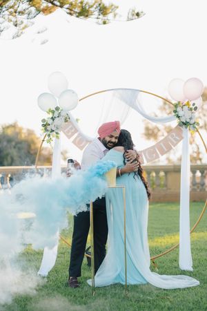 A romantic outdoor wedding moment under a decorated arch with blue smoke and floral accents in natural sunlight.