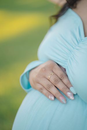 A close-up of a pregnant woman's belly in a light turquoise dress, showing her hand with French manicure and engagement ring.