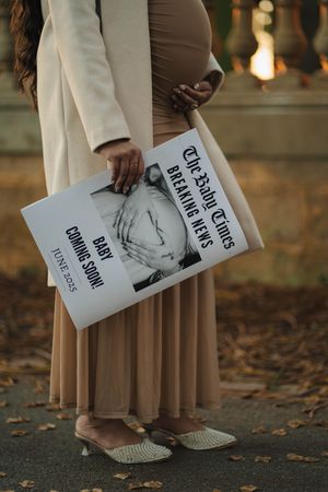 A pregnant woman in a tan dress holding an ultrasound book stands on autumn leaves wearing casual slip-on shoes.