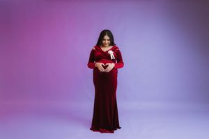 Figure in a long red velvet dress holds book while standing against purple backdrop in studio setting.