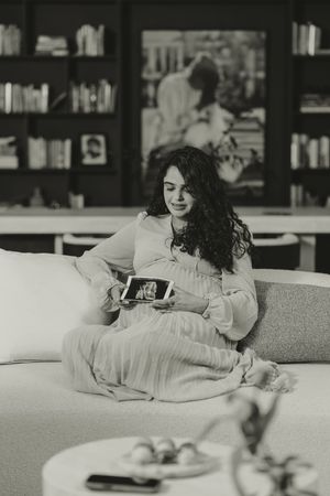 Pregnant woman sitting on sofa holding ultrasound photo, black and white portrait with bookshelves in background.