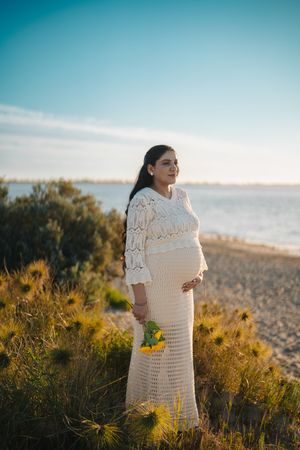 A pregnant woman in a white lace dress stands on a coastal cliff at sunset holding yellow flowers.