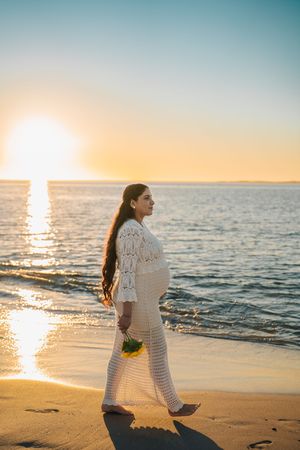 A person in a white crochet dress walks along a sandy beach at sunset while ocean waves roll in.