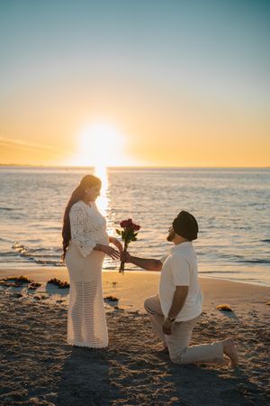 A romantic beach proposal at sunset with silhouettes kneeling in the sand by the ocean.
