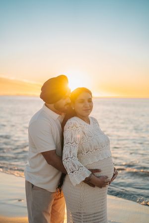 Couple embraces on beach at sunset while expecting a baby, with woman wearing white lace dress against golden sky.