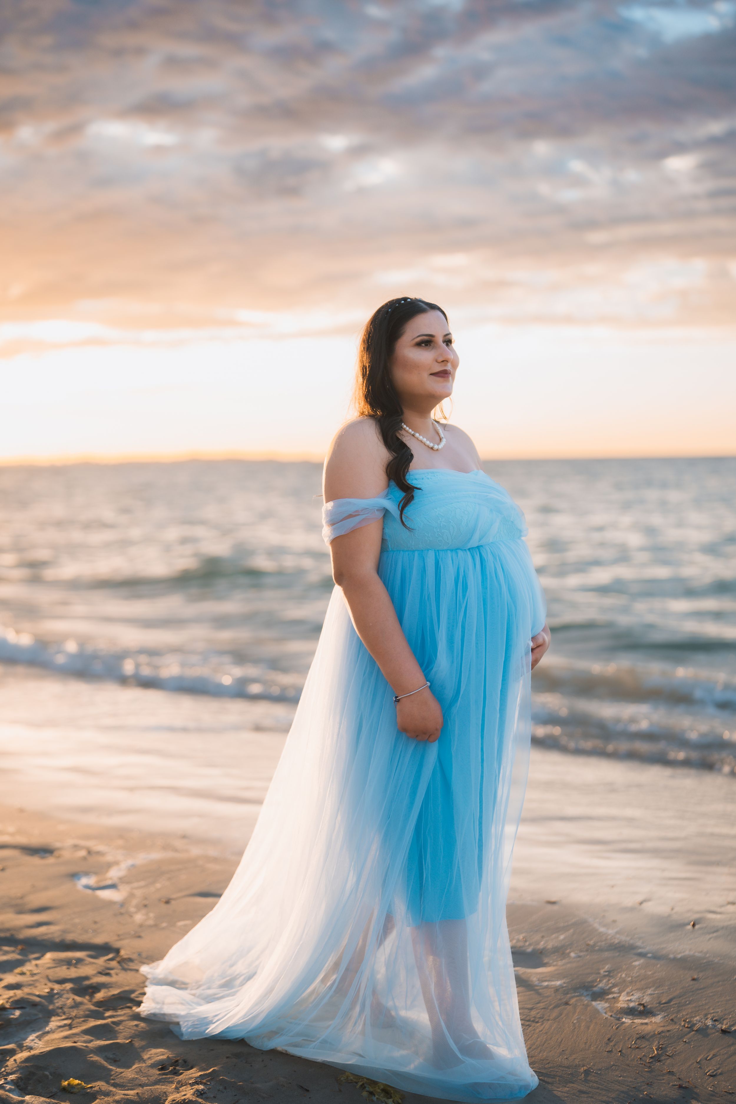 A person in a flowing blue and white dress poses gracefully on a beach at sunset creating a dreamy coastal scene.