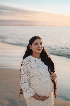 A person in a white lace dress poses at sunset on a beach with ocean waves in the background.
